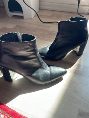 Svarta boots med klack och dragkedja - Säljer ett par svarta stövletter i skinnimitation med bred klack och dragkedja på sidan. De har en spetsig tå och stilren design som passar till många olika outfits. Perfekta för dig som vill ha en edgy men klassisk look.