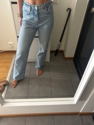 Ljusblå raka jeans med hög midja från Zara - Säljer ett par ljusblå jeans med rak passform och hög midja. Jeansen har klassiska fem fickor, bälteshällor och stängs med knapp och dragkedja. Perfekta till en avslappnad och trendig look.
