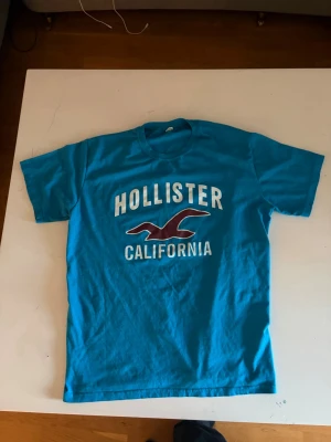 Blå Hollister t-shirt med tryck - Snygg blå t-shirt från Hollister med stort vitt och vinrött tryck på bröstet där det står 'Hollister California' och en ikonisk fågel. Klassisk passform och rund hals. Perfekt till jeans eller shorts. Knappast använd. ABS! Pris går att diskutera.