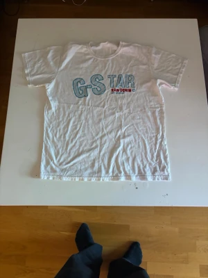 Vit G-Star RAW t-shirt med tryck - Vit t-shirt från G-Star RAW med stort blått och rött tryck på bröstet där det står 'G-STAR RAW DENIM'. Klassisk passform med rund hals och korta ärmar. Perfekt för en avslappnad och stilren look. Knappast använd. ABS! Pris går att diskutera.