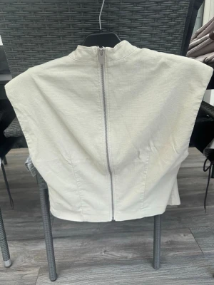 Vit ärmlös väst med dragkedja - Stilren vit väst i ett strukturerat tyg med hög krage och markerade axlar. Västen är ärmlös och har en dragkedja framtill som ger en modern look. Perfekt att styla över en skjorta eller t-shirt för en trendig outfit.