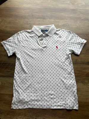  Polo Ralph Lauren - Vit tröja från Polo Ralph Lauren med små mörkblå segelbåtar som mönster och den klassiska röda loggan broderad på bröstet. Tröjan har korta ärmar, krage och knappar framtill. Perfekt för en avslappnad men snygg stil. Slim fit
