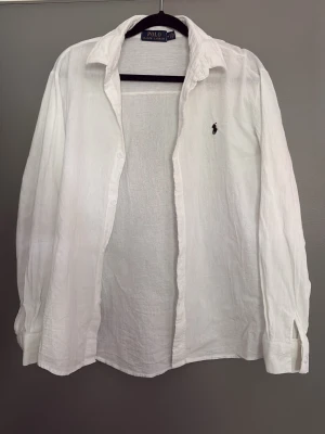 Vit skjorta från Polo Ralph Lauren - Klassisk vit skjorta i linne från Polo Ralph Lauren med broderad logga på bröstet. Skjortan har lång ärm, knappar framtill och en avslappnad passform. Perfekt till sommaren för en fräsch och stilren look.