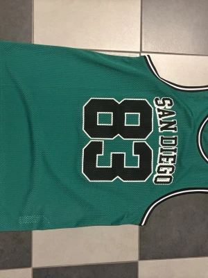 Grön basketlinne med San Diego-tryck - Säljer ett grönt basketlinne med svartvitt tryck där det står 'San Diego' och siffran 83 på ryggen. Linnet har vita och svarta detaljer vid halsringning och ärmhål. Tillverkat i meshmaterial som andas, perfekt för träning eller streetwear.