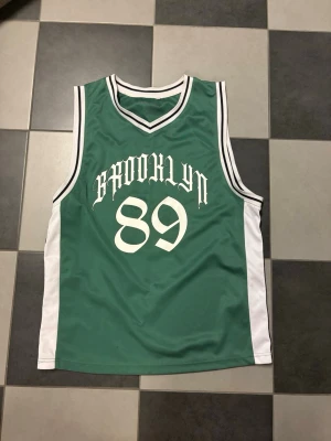 Grön basketlinne med BROOKLYN 89 - Snyggt grönt basketlinne med vita sidopaneler och svartvita ränder vid hals och ärmhål. Tryck med texten 'BROOKLYN' och siffran 89 på bröstet. Tillverkad i ett lätt och ventilerande material, perfekt för träning eller streetwear.