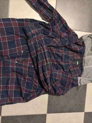 Rutig overshirt från Vans med huva - Snygg overshirt från Vans i blått och vinrött rutmönster. Skjortan har grå huva i sweatshirtmaterial och knäppning framtill. Perfekt lager på lager-plagg med casual streetkänsla och mjukt tyg.