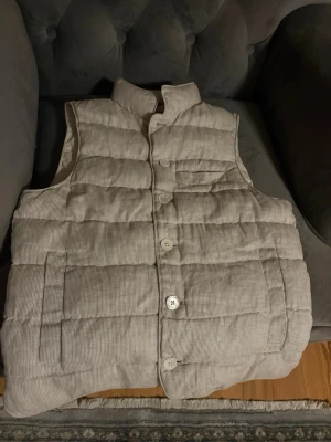 Beige ribbad dunväst med knappar - Använt en gång passar i storlek S men även M. Säljer en beige dunväst med ribbad struktur och stora vita knappar framtill. Västen har hög krage, två sidofickor och en bröstficka. Perfekt för lager på lager och ger en chill look till din outfit. 