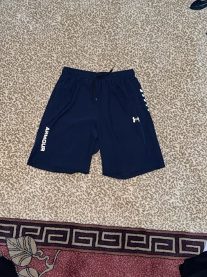 Marinblå träningsshorts Under Armour - Säljer ett par marinblå träningsshorts från Under Armour med elastisk midja och snörning. Shortsen har sidofickor med dragkedja och färgglada detaljer längs sidan samt logga på benet. Perfekta för gymmet eller löprundan.