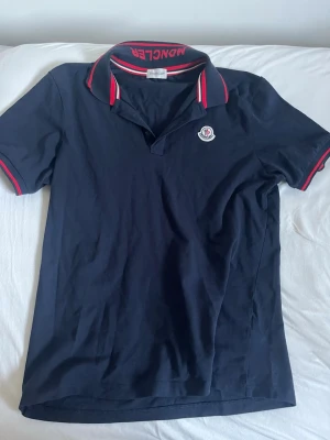 Marinblå pikétröja från Moncler - Snygg marinblå pikétröja från Moncler med klassisk krage och röda samt vita detaljer på krage och ärmslut. Tröjan har Moncler-logga på bröstet och text på insidan av kragen. Perfekt för en stilren och sportig look.