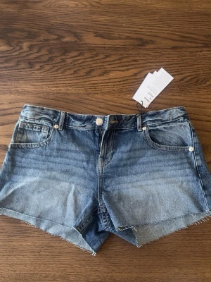 Blå jeansshorts från Gina Tricot - Säljer ett par klassiska blå jeansshorts från Gina Tricot. De har råa kanter, fem fickor och knappgylf. Perfekta för sommaren och har en avslappnad passform med hög midja. Snygg tvättad denim-look som passar till allt.