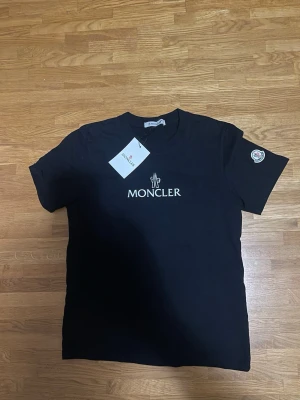 Svart Moncler t-shirt med logga - Svart t-shirt från Moncler med vit logga tryckt på bröstet och ett Moncler-märke på ärmen. Klassisk rund hals och korta ärmar. Tillverkad i mjuk bomull för en skön känsla. Perfekt för dig som gillar stilrena och exklusiva plagg.