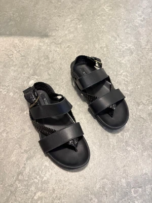 Svarta sandaler från Tiger of Sweden - Snygga svarta sandaler från Tiger of Sweden med breda remmar i läder och detaljer i flätad design. Justerbart spänne vid ankeln och öppen tå. Perfekta för en trendig och avslappnad look.