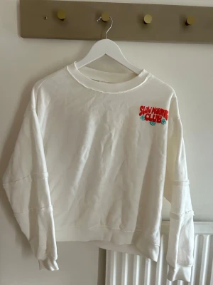 Vit sweatshirt - Vit sweatshirt med tryck' i rött och turkost på bröstet och stor grafik på ryggen med frukostmotiv. Tröjan har rund hals, långa ärmar och ribbade muddar. Perfekt för chill vibes och avslappnade dagar.