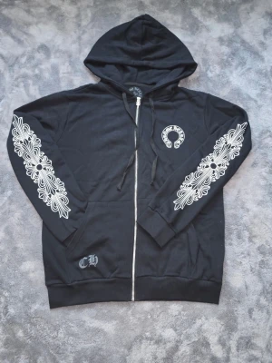 Svart Chrome Hearts zip hoodie - Svart hoodie med dragkedja från Chrome Hearts. Stora vita tryck på ärmarna och ryggen, samt mindre logga på bröstet. Huva med dragsko och ribbade muddar. Materialet är mjukt och bekvämt, perfekt för en streetwear-look.