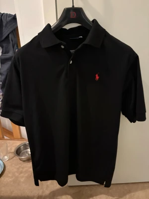 Ralph lauren Svart pikétröja med röd logga - Klassisk svart pikétröja med korta ärmar och  krage. Tröjan har två knappar framtill och en liten röd broderad logga på bröstet. Perfekt till jeans eller shorts för en enkel och stilren look.