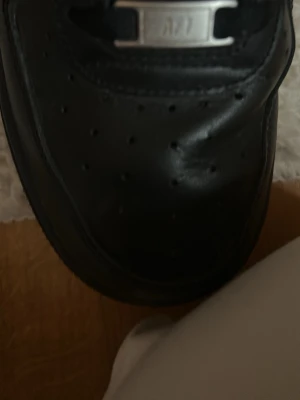 Svarta Nike Air Force 1 sneakers - Klassiska helsvarta Nike Air Force 1 sneakers i läder med perforerad tåbox och svarta skosnören. Snygg metallbricka med 'AF1' på snörningen och diskret Nike Air-logga på plösen. Perfekt för dig som gillar tidlös streetstil.