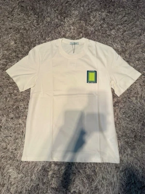 Vit t-shirt med grafiskt tryck - Vit t-shirt med rund hals och korta ärmar. På bröstet finns ett litet grafiskt tryck i grönt och blått, och på ryggen ett större tryck med texten 'Casa Blanca' och färgade rektanglar. Perfekt för en trendig och avslappnad stil.