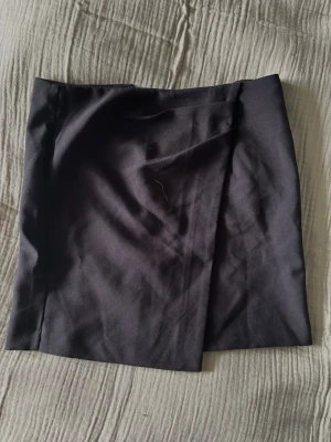 Svart omlottkjol från BikBok - Snygg svart kort omlottkjol från BikBok i stilren design. Kjolen har en diskret omlott-detalj framtill och stängs med dragkedja i sidan. Perfekt att matcha med en crop top eller stickad tröja. Helt ny🌸