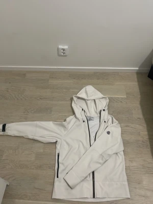 Vit vindjacka från Moncler - Snygg vit vindjacka från Moncler med svart dragkedja, två sidofickor med dragkedja och justerbar huva. Jackan har en stilren design och Moncler-logga på ärmen. Perfekt för blåsiga dagar och enkel att matcha med olika outfits.