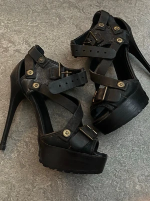 Svarta platåpumps med nitar och spännen - Säljer ett par svarta pumps med hög platå och stilettklack. Skorna har flera remmar med metallspännen och dekorativa nitar i mässing. Öppen tå och robust sula ger en edgy look. Perfekta för dig som gillar statementskor och vill sticka ut.