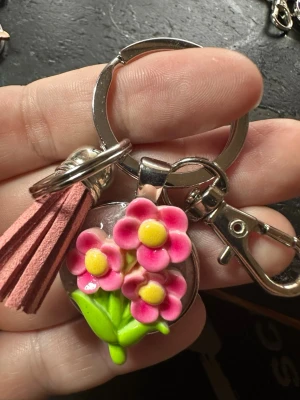 Nyckelring med rosa blommor och tofs - Söt nyckelring med en dekoration av tre rosa blommor med gula mittpunkter och gröna blad i plast. Nyckelringen har även en rosa tofs i mockaimitation och en metallhake för enkel fastsättning. Perfekt accessoar för att piffa upp dina nycklar eller väska.