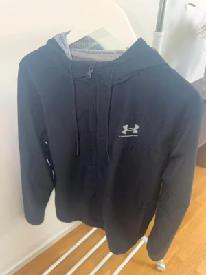  Under Armour - Snygg svart vindjacka från Under Armour med huva och halv dragkedja framtill. Jackan har justerbar dragsko i huvan, elastiska muddar vid ärmsluten och ett diskret logotryck på bröstet. Tillverkad i lätt och vindtätt material, perfekt för blåsiga dagar. Storlek L