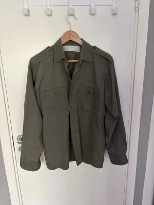 Olivgrön overshirt med fickor - Säljer en olivgrön overshirt med två bröstfickor med knappar och axelklaffar. Overshirten är använd fåtal gånger och har inga defekter eller liknande, hör av er vid frågor!