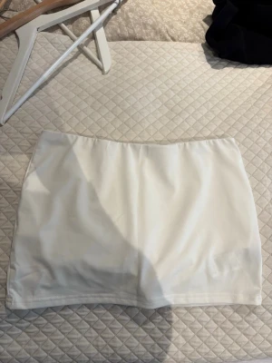 Vit kortkjol i syntetmaterial - Stilren vit kortkjol med enkel och rak passform. Kjolen är gjord i ett slätt syntetmaterial och har inga synliga detaljer som fickor eller dragkedja. Perfekt att matcha med olika toppar för en clean look.