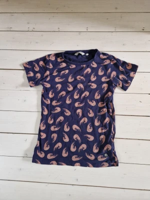 Mörkblå t-shirt med räkmönster - Säljer en mörkblå t-shirt från EmmaMalena med korta ärmar och ett unikt mönster av ljusrosa räkor över hela tröjan. Tillverkad i mjuk bomull, perfekt för dig som gillar lekfulla prints och vill sticka ut lite extra.