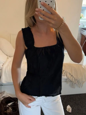 Svart linnetopp med breda axelband - Snygg svart linnetopp med breda axelband och rak halsringning. Toppen har en enkel och stilren design som passar perfekt till både jeans och kjol. Materialet ser ut att vara bomull eller linne för en luftig känsla.