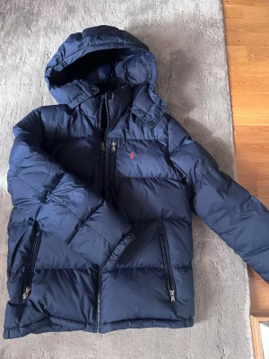 Marinblå dunjacka från Polo Ralph Lauren - Snygg marinblå dunjacka från Polo Ralph Lauren med huva och dragkedjor på fickorna. Jackan har en klassisk pufferstil och den ikoniska röda loggan på bröstet. Perfekt för den kommande vinter säsongen och enkel att matcha med olika outfits.