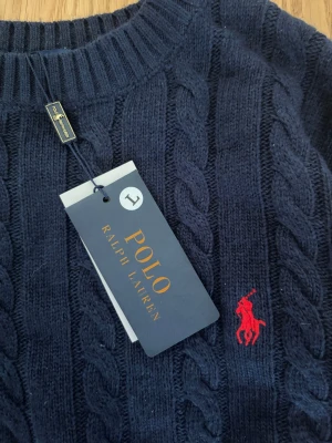 Mörkblå kabelstickad tröja Polo Ralph Lauren - Kabelstickad tröja i mörkblått från Polo Ralph Lauren med rund halsringning och den klassiska röda logotypen broderad på bröstet. Tröjan har långa ärmar och ribbade muddar vid ärmslut och nederkant. Perfekt för en stilren och avslappnad look. Obs. (Tröjan är storlek L men sitter mer som en M) Aldrig använd hör av dig vid frågor.