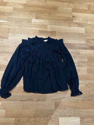 Marinblå blus med volanger och glitter - Supersnygg marinblå blus med långa ärmar, volangdetaljer och diskreta glittriga trådar i tyget. Blusen har hög krage med rysch och smock upptill, vilket ger en romantisk vibe. Perfekt att styla med jeans eller kjol för en trendig look. Också perfect för speciella tillfällen.