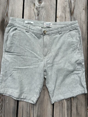 Ljusgråa linneshorts med fickor - Snygga ljusgrå shorts i linneblandning med klassisk passform. Shortsen har sidofickor fram, två bakfickor med knapp och bälteshällor i midjan. Perfekta för varma dagar och ger en clean och stilren look.