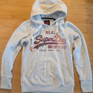 Ljusgrå hoodie från Superdry - Sååå fin hoodie från superdry med rosa detaljer!! Storlek xs.  Jätte fint skick! Säljer pga att jag har andra Hoodies som jag föredrar! Sällsynt hoodie som inte går att köpa längre tror jag, hör av dig vid funderingar!🩷
