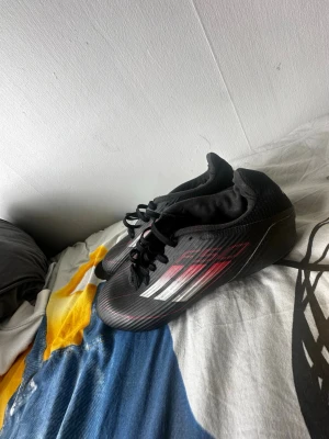 Svarta fotbollsskor med rödvit detalj - Säljer ett par svarta fotbollsskor med snygga röda och vita detaljer på sidan. Skorna har snörning och låg profil, perfekta för snabba vändningar på planen. Yttermaterialet är syntetiskt och designen är sportig och modern.