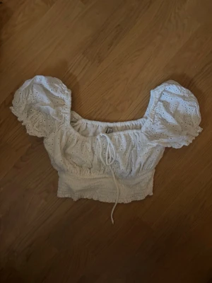 Vit blus med puffärmar och spetsdetaljer - Supersöt vit blus med puffiga kortärmar och broderade spetsdetaljer. Toppen har smockad midja och knyt framtill för extra söt look. Perfekt till jeans eller kjol för en trendig sommarstil.