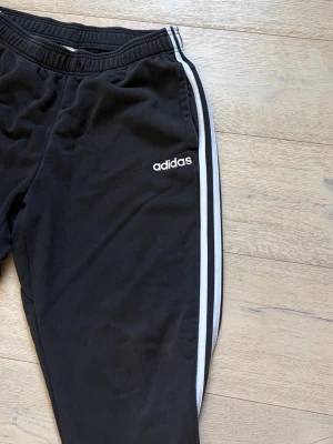 Svarta Adidas mjukisbyxor med ränder - Säljer ett par svarta mjukisbyxor från Adidas med klassiska vita ränder längs sidorna och Adidas-logga på låret. Byxorna har resår i midjan och muddar vid bensluten. Perfekta för chill eller träning.