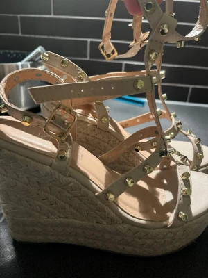Beige kilklackade sandaler med nitar - Säljer ett par beige sandaler med hög kilklack i flätad jute och flera remmar dekorerade med guldfärgade nitar. Sandalerna har justerbara spännen och öppen tå. Perfekta för dig som vill ha en edgy men ändå somrig look.