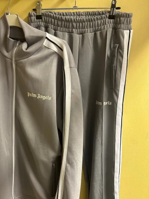 Grå Palm Angels tracksuit, M - Säljer en grå tracksuit från Palm Angels med vita och svarta ränder längs ärmar och ben. Setet består av zip-jacka och matchande byxor med elastisk midja och sidofickor. Palm Angels-logga broderad på både jacka och byxa. Perfekt för chill eller streetwear. Helt nya. 