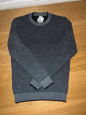 Grå stickad tröja från DeFacto - Snygg grå stickad tröja från DeFacto med rund halsringning och diskret rutmönster. Tröjan har ribbade muddar vid ärmslut och nederkant samt en klassisk standard fit. Perfekt till höstens och vinterns outfits.