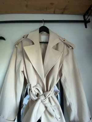 Beige trenchcoat med knytskärp - Stilren beige trenchcoat med breda slag, axelklaffar och knytskärp i midjan. Jackan har en klassisk siluett och är tillverkad i ett slätt tyg som ger en elegant look. Perfekt för dig som vill ha en tidlös och trendig ytterjacka.