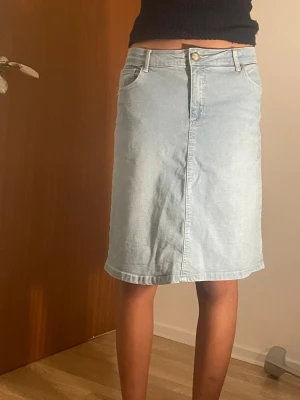 Ljusblå jeanskjol med fickor - Säljer en ljusblå jeanskjol i klassisk modell med fem fickor och knappgylf. Kjolen har en rak passform och går till knäna. Perfekt till vardags för en avslappnad och trendig look. Knappt använd! 
