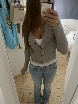 Beige ribbad kofta med knappar - Säljer en beige ribbad kofta med djup v-ringning och stora bruna knappar framtill. Koftan är croppad och har långa ärmar. Perfekt att slänga över ett linne eller t-shirt för en chill look.