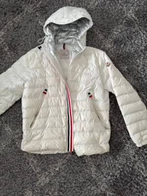 Vit dunjacka med huva från Moncler - Snygg vit dunjacka från Moncler med glansig finish och silvrig insida på huvan. Jackan har två dragkedjefickor, Moncler-logga på ärmen och detaljer i rött, vitt och blått vid dragkedjan och fickorna. Perfekt för kalla vinterdagar.