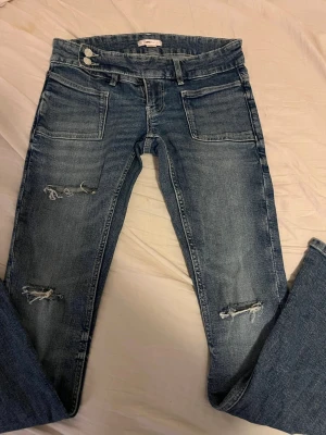 Blå slitna jeans från Gina Tricot - Säljer ett par blå jeans från Gina Tricot med slitningar på båda benen och raka ben. Jeansen har stora framfickor, dubbla knappar i midjan och är tillverkade i ett klassiskt denimtyg. Perfekta för en avslappnad och trendig look. Har använts kanske två gånger så de är i princip som nya! 
