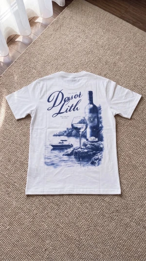 Vit t-shirt med blått vintagemotiv - Vit t-shirt med stort blått tryck på ryggen som föreställer en vinflaska, ett glas och en havsmiljö. Texten 'Dosoi Lith' och dekorativa detaljer ger en vintagekänsla. Klassisk passform och rund hals, perfekt för en avslappnad stil.