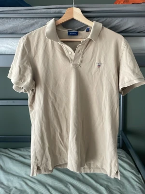 Beige pikétopp från GANT - Beige pikétopp från GANT med klassisk krage, korta ärmar och knappar framtill. Liten broderad logga på bröstet. Tillverkad i mjuk bomull för en clean och avslappnad stil.