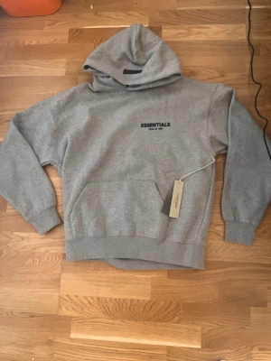 Grå Essentials hoodie med tryck - Snygg grå hoodie från Essentials med svart tryck 'ESSENTIALS Fear of God' på bröstet och ryggen. Klassisk känguruficka framtill och stor huva. Perfekt för en chill och avslappnad stil. Mjuk bomullsblandning för extra komfort.