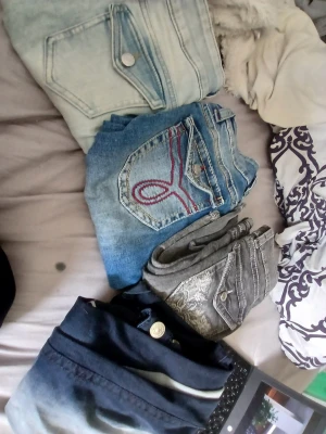 Fyra par jeans i olika färger - Säljer ett paket med fyra par jeans i olika tvättar: ett par ljusblå, ett par klassiskt blå med broderad bakficka, ett par grå med dekorativ söm och ett par mörkblå. Perfekt för dig som vill variera din stil med olika färger och detaljer.